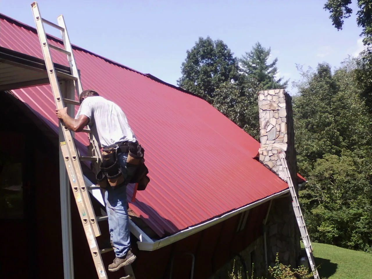 Roofer on a ladder beside a red metal roof and stone chimney — Soffit & Fascia Repair by Roofing Brothers in New Matamoras